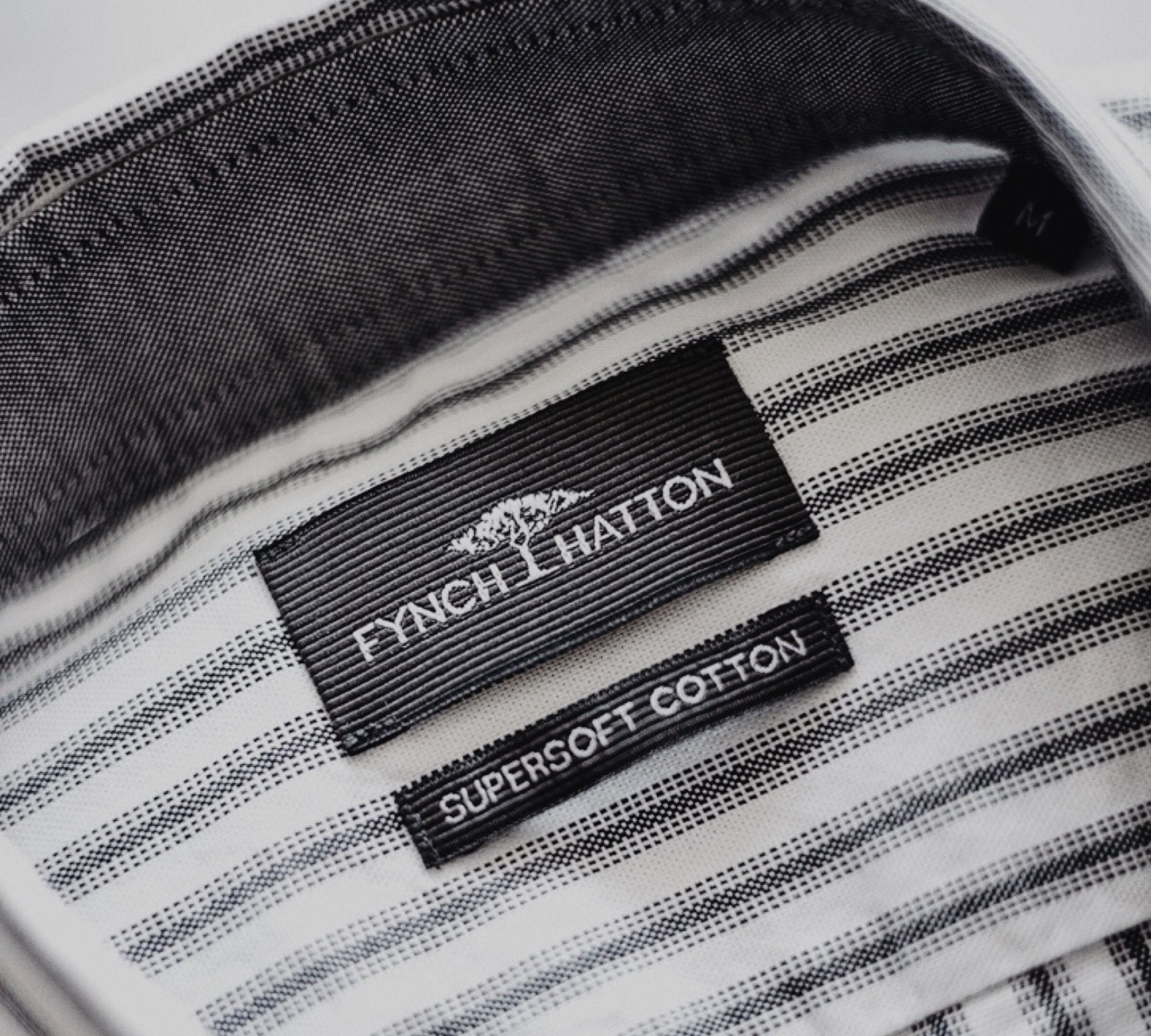 Innenansicht eines gestreiften Hemdkragens mit zwei schwarzen Etiketten – eines mit Markenname FYNCH-HATTON, das andere mit der Aufschrift SUPERSOFT COTTON – Text: Produkt & Qualität.
